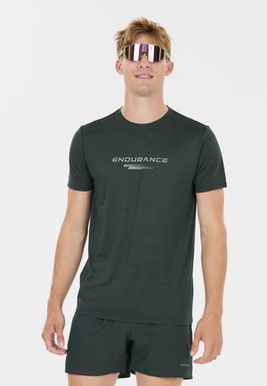 Jonge man in een zwart "Endurance" T-shirt, bijpassende korte broek en reflecterende sportzonnebril, staand tegen een effen witte achtergrond.