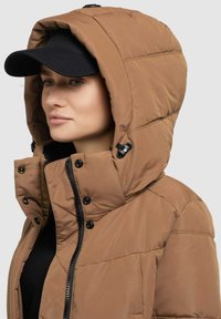 Braune Pufferjacke mit Kapuze, mit gestepptem Muster, schwarzem Zubehör und Reißverschluss. Eine schwarze Mütze rundet den Look ab.