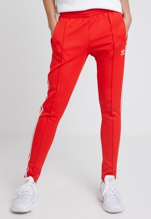 Personne portant un pantalon de survêtement rouge Adidas avec des bandes blanches et des baskets blanches, debout les mains dans les poches devant un fond uni.