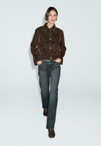 Kvinde går iført en brun ruskindsjakke med klaplommer, lige ben jeans i blå, et naglet bælte og brune loafers mod en ensfarvet baggrund.
