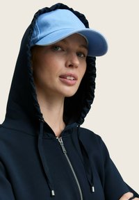 Žena u tamnoplavoj dukserici s patentnim zatvaračem i kapuljačom, s laganom svijetloplavom bejzbol kapom, gleda lagano u stranu pred jednostavnom pozadinom.