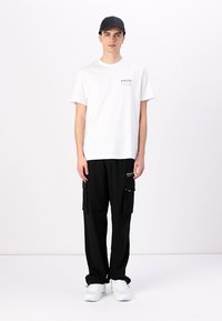 Witte katoenen t-shirt met een klein logo, gecombineerd met zwarte cargo broek met zijzakken en hardwaredetails. Accessoires bestaan uit een zwarte pet.
