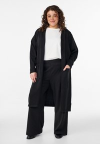 Schwarzer langer Mantel mit offenem Vorderteil, ausgestattet mit zwei Vordertaschen. Getragen über einem weißen Oberteil und schwarzen Hosen, strukturierter Stoff. Modell lächelt.