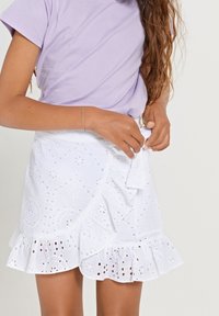 Bílé sukně z očkovaného materiálu s volánkovým lemem, která obsahuje složité výřezy a detail vázání v pase. Bavlněný materiál a měkká textura.