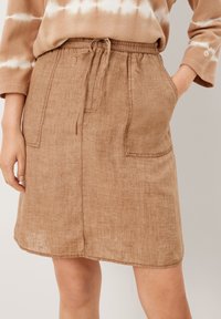 Donna che indossa una gonna di lino marrone fino al ginocchio con tasche applicate e vita regolabile tramite cordoncino, abbinata a una maglietta beige con motivo tie-dye.