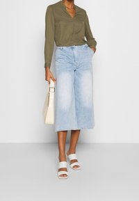 Zielona oliwkowa bluzka z długim rękawem, jasnoniebieskie szerokie dżinsowe kuloty, białe sandały na obcasie oraz torebka w kolorze kremowym o eleganckim designie.