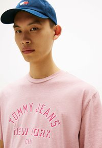 Roze katoenen T-shirt met "TOMMY JEANS NEW YORK" in het wit en rood gedrukt, gecombineerd met een blauwe pet met een logo patch.