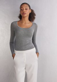 Grijze long-sleeve shirt met een ronde hals, gecombineerd met witte broeken met een platte tailleband en zijzakken, geplaatst tegen een neutrale achtergrond.