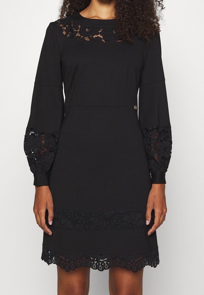 Femme portant une robe noire à manches longues avec des détails en dentelle sur le décolleté, les manches, la taille et l'ourlet.