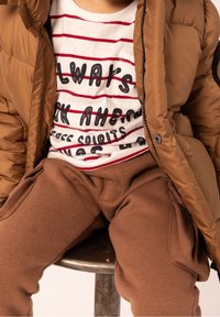 Braune gepolsterte Jacke mit Reißverschluss, über einem weißen T-Shirt mit roten Streifen und schwarzem Text getragen. Braune Jogginghose mit Seitentaschen.