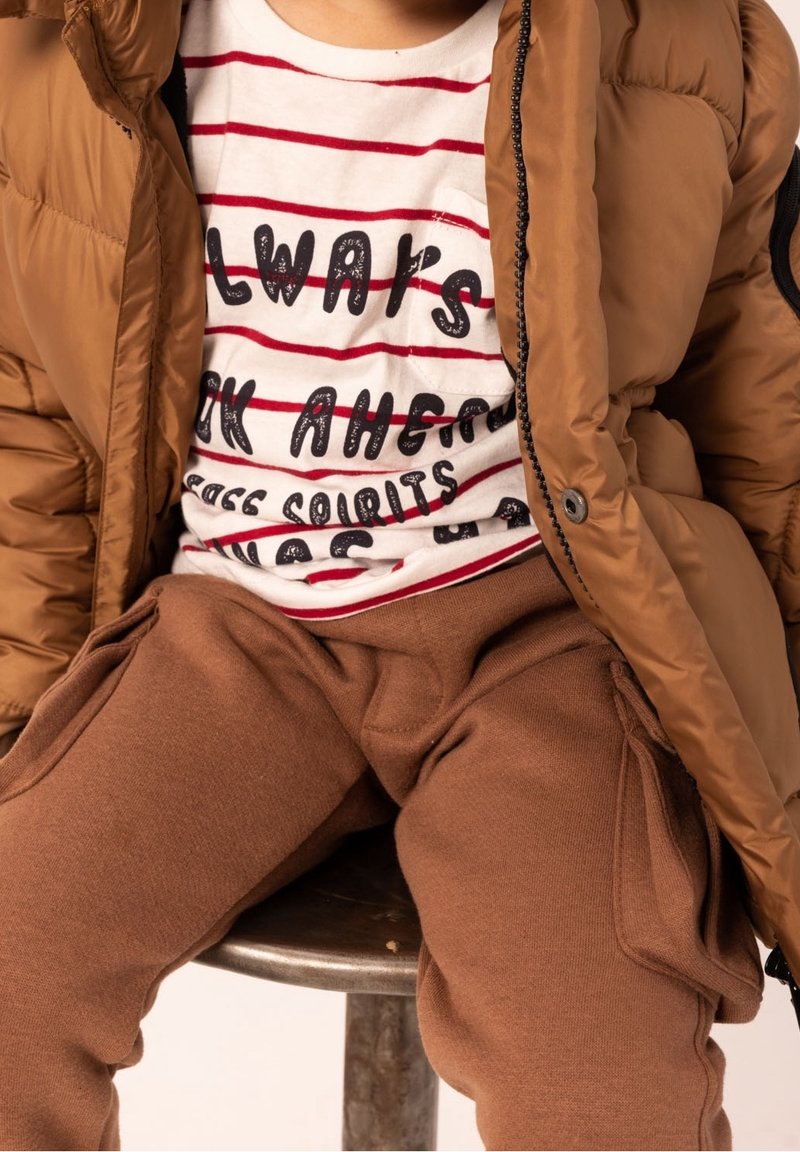Braune gepolsterte Jacke mit Reißverschluss, über einem weißen T-Shirt mit roten Streifen und schwarzem Text getragen. Braune Jogginghose mit Seitentaschen.