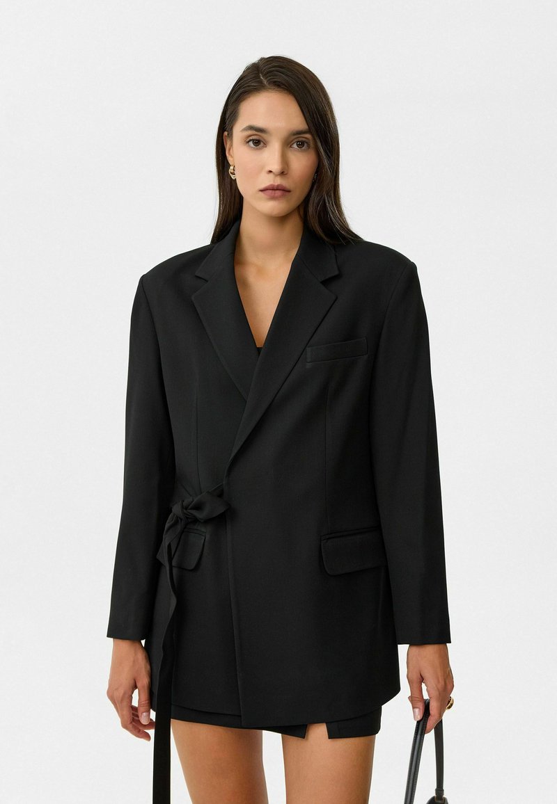 Zwarte blazer met een strik in de taille, enkele borstzak, lange mouwen en gestructureerde schouders. Gladde stof met een matte afwerking.