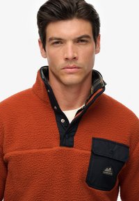 Fleece pullover in burnt orange with a quilted black collar, snap closure, and a chest pocket featuring a logo patch. Textured and warm.