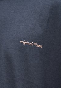 Σκούρο γκρι ύφασμα με μικρό ανοιχτό ροζ κείμενο που γράφει «originals © 1990» στο κέντρο.