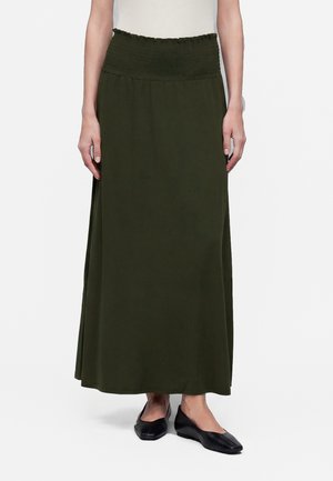 Femme portant une longue jupe vert olive avec une taille froncée et des chaussures plates noires, debout devant un fond blanc.
