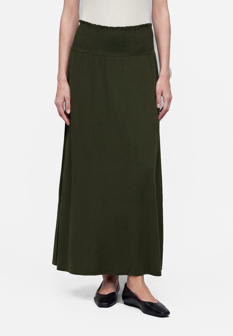 Femme portant une longue jupe vert olive avec une taille froncée et des chaussures plates noires, debout devant un fond blanc.