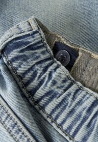 Tela de mezclilla con un patrón texturizado en azul y blanco, que presenta un cierre de botón y costuras detalladas sobre un fondo azul claro.