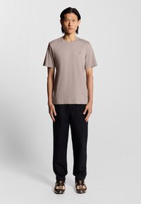 T-shirt a maniche corte marrone chiaro con piccolo logo, abbinato a pantaloni neri e sneaker marroni con motivo. Design semplice e rilassato.