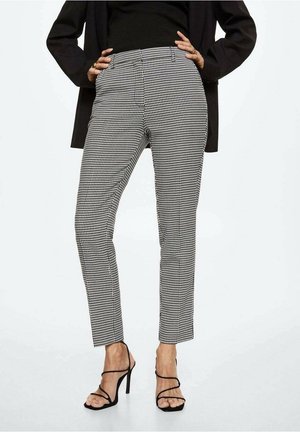 Vrouw die zwarte en witte slanke broek met ruitpatroon draagt, zwarte blazer en zwarte sandalen met hoge hakken en bandjes, handen in de zij.