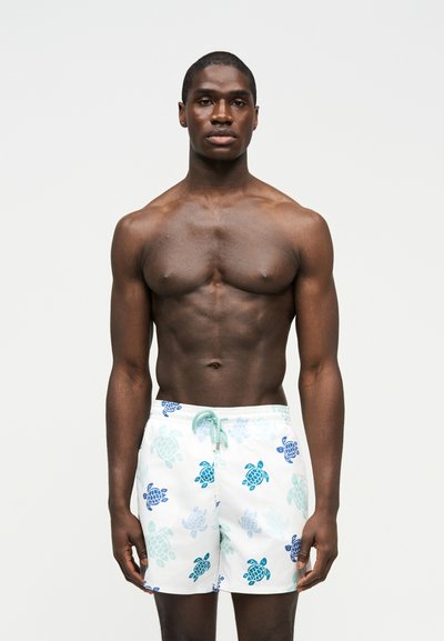 Μυώδης άνδρας που φοράει λευκά μαγιό με μπλε και τιρκουάζ σχέδια θαλάσσιων χελωνών σε απλό ανοιχτό φόντο.