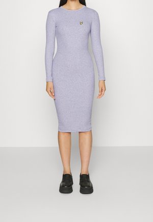 Femme portant une robe midi lavande ajustée à manches longues avec un petit logo d'oiseau jaune sur la poitrine et des chaussures plateformes noires.