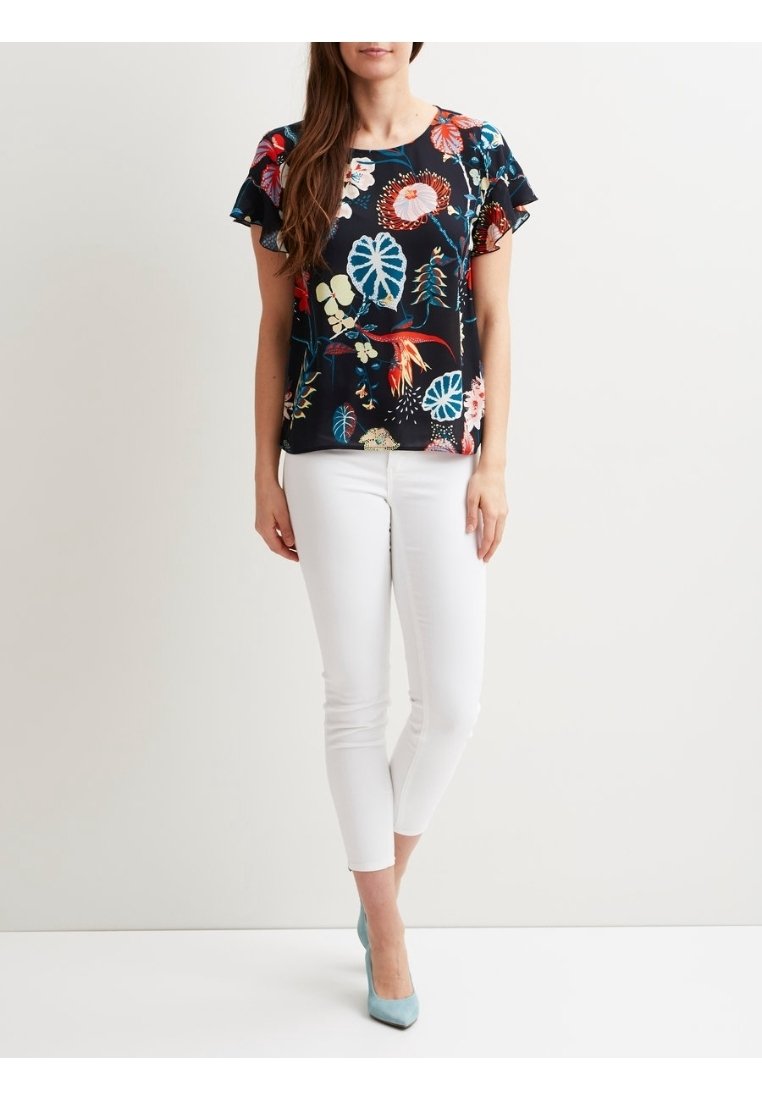 Femme portant un chemisier à manches courtes à fleurs foncées, un pantalon blanc ajusté et des chaussures à talons hauts bleu clair, debout sur un fond uni.