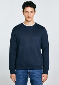 Jonge man met kort donker haar, gekleed in een marineblauwe trui en een blauwe spijkerbroek, staand tegen een effen lichte achtergrond.