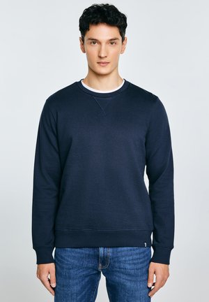 Noor mees lühikeste tumedate juustega, seljas tumesinist värvi dressipluus ja sinised teksad, seisab ühevärvilise hele tausta ees.