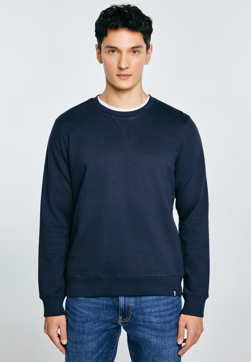 Jonge man met kort donker haar, gekleed in een marineblauwe trui en een blauwe spijkerbroek, staand tegen een effen lichte achtergrond.