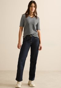 Top grigio a maniche corte con scollatura a V, abbinato a jeans dritti blu scuro e sneaker bianche, su uno sfondo neutro.