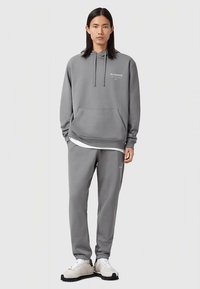 Vīriešu modelis ar gariem tumšiem matiem, tērpies pelēkā kapucē un atbilstošos sporta biksēs, baltās kedās, stāvot uz viendabīga gaiša fona.