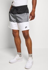 Muž oblečený v černých, šedých a bílých Nike šortkách s šedými Nike ponožkami a bílými teniskami, stojící na jednobarevném bílém pozadí.