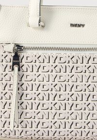 Balta DKNY rankinė, kurioje matyti firminė oda, metalinis užtrauktukas su odine kilpele ir pasikartojantis DKNY logotipo raštas ant audinio.