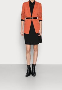 Oranžový blazer s černým páskem, vrstvený přes černé šaty. Tento outfit má na míru ušitý design, strukturované ramena a elegantní černé kotníkové boty.