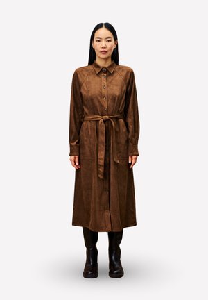 Femme se tenant de face portant une robe midi marron à boutons avec une ceinture nouée à la taille et des bottes noires hauteur genou sur un fond blanc.