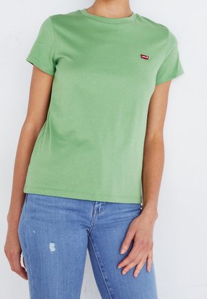 Donna che indossa una t-shirt Levi's verde chiaro a maniche corte e jeans strappati azzurri chiaro su uno sfondo bianco semplice.