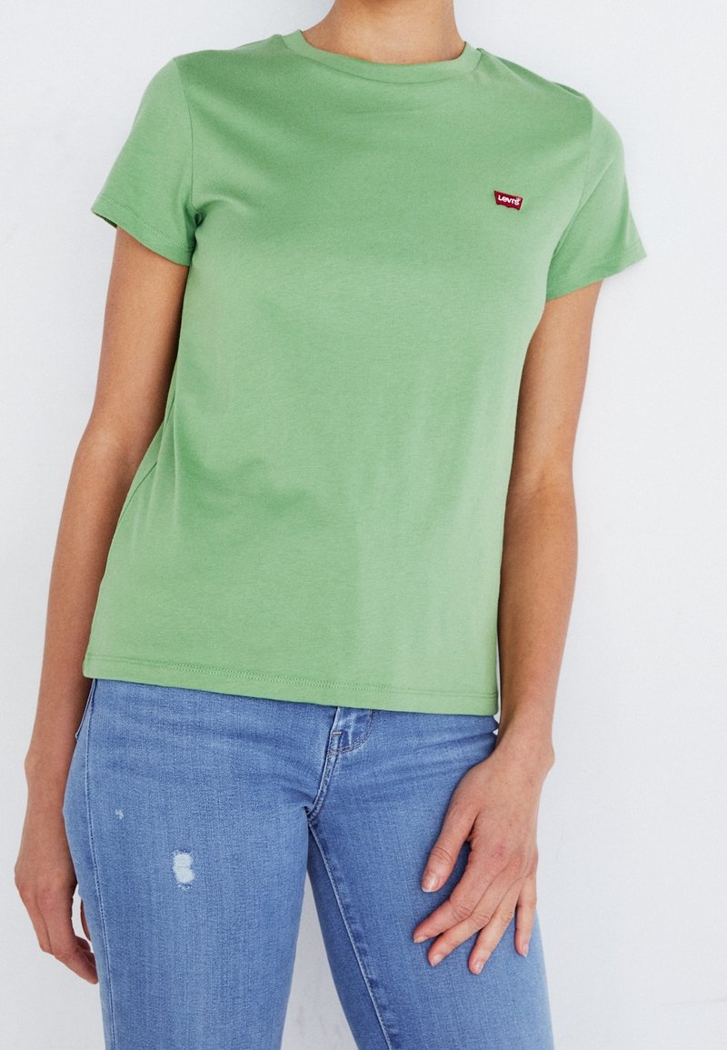 Femme portant un t-shirt Levi's vert clair à manches courtes et un jean bleu clair déchiré, sur fond blanc uni.