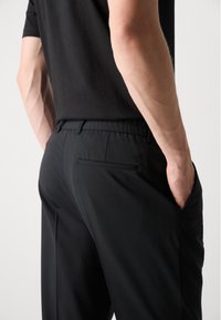 Pantaloni negri cu o croială lejeră, având un elastic în talie și un singur buzunar pe spate. Materialul pare neted, cu o textură minimă.
