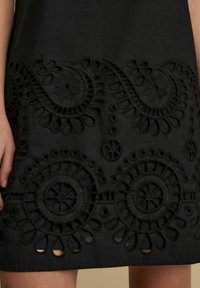 Schwarzes Kleider aus Stoff mit aufwendigen bestickten Details in floralen und kreisförmigen Mustern, das ausgeschnittene Akzente am Saum zeigt.