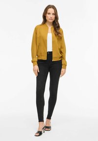 Veste bomber moutarde à silhouette ajustée, poignets côtelés, détails importants de fermeture éclair, assortie à un jean skinny noir taille haute et des talons.