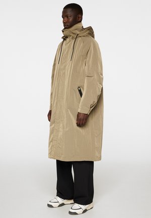 Manteau de pluie long beige avec une finition texturée, capuche ajustable, poches avant et cordons de serrage noirs, superposé sur un pantalon noir et des chaussures.