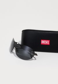 Juodieji Diesel saulės akiniai su elegantišku, lenktu objektyvu ir sidabriniais aksesuarais. Komplekte yra juodas dėklas su raudonu Diesel logotipu.