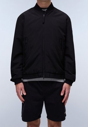 Mężczyzna noszący czarną kurtkę bomber na zamek błyskawiczny z prążkowanymi mankietami i dołem, czarne spodenki oraz szarą koszulę pod spodem.