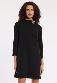 Robe noire à manches trois-quarts, avec un col montant et un accent floral doré sur l'épaule. Texture lisse, coupe droite.