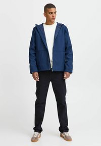 Veste zippée bleu marine avec capuche, dotée de deux poches avant et d'une texture de tissu lisse. Portée par-dessus une chemise blanche et un jean noir.