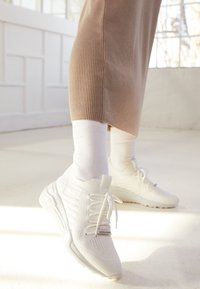 White athletic sneakers made of mesh and synthetic materials, featuring a textured pattern, laces, and a cushioned sole. Worn with white socks.