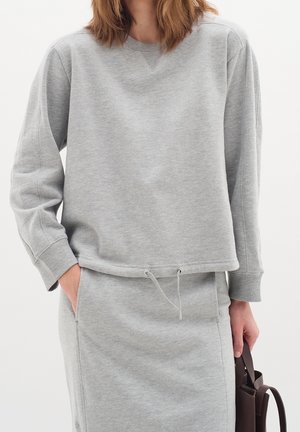 Femme portant un sweat-shirt gris clair et une jupe assortie, tenant un sac à main marron foncé, une main dans la poche.