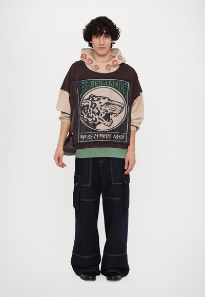 Hoodie graphique marron, beige et vert avec un design de tigre et du texte, associé à un pantalon cargo en denim foncé ample avec des poches latérales.