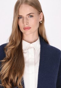 Tmavomodrý kardigán s textúrovaným pletením, nosený cez bielu košeľu na gombíky. Modelka má dlhé, rovné vlasy a kryštálové náušnice.