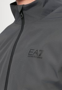 Prim-plan al unei jachete gri închis, cu fermoar frontal și logo-ul negru "EA7 Emporio Armani" pe piept.